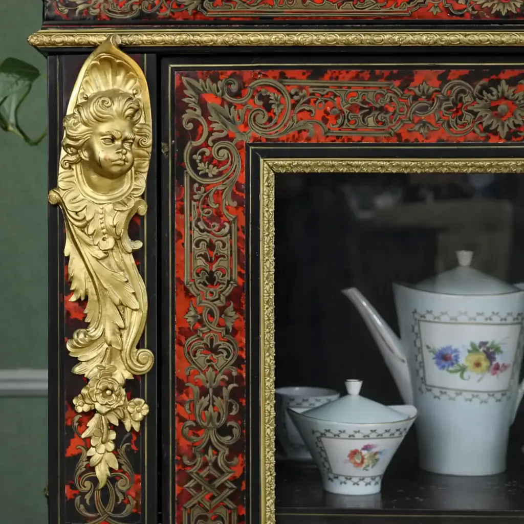 Vetrina alla Boulle in ebano, tartaruga e bronzi dorati Francia, seconda metà del XIX secolo – Stile Napoleone III Raffinata vetrina a un’anta realizzata secondo la celebre tecnica “alla Boulle”, con impiallacciatura in ebano e ricchi intarsi in ottone su fondo in tartaruga rossa naturale, disposti in eleganti motivi a racemi e volute di gusto Luigi XIV. Il prospetto è impreziosito da importanti applicazioni in bronzo dorato finemente cesellato, raffiguranti figure allegoriche laterali, fregi a foglie d’acanto e mascherone centrale nella parte inferiore. L’anta vetrata, incorniciata da profili dorati, apre su ripiani interni destinati all’esposizione di porcellane e oggetti d’arte. Esempio di alta ebanisteria francese del periodo Napoleone III, il mobile coniuga ricchezza ornamentale, equilibrio architettonico e forte impatto scenografico, risultando ideale per ambienti di rappresentanza o collezioni antiquarie di pregio.