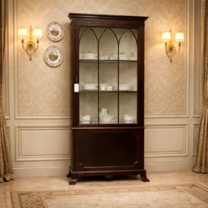 Angoliera a due corpi in legno massello, stile inglese, fine XIX secolo Elegante angoliera di fine Ottocento in legno massello con finitura a gommalacca color mogano scuro, impostata su pianta triangolare per collocazione ad angolo. Il mobile è composto da un corpo unico: la parte superiore con anta vetrata scandita da traverse ad arco ogivale, di gusto tardo vittoriano, destinato all’esposizione; l’inferiore con anta cieca a pannello sagomato. Interni con ripiani in legno e rivestimento decorativo, base su piedi sagomati a mensola. Linee sobrie ed equilibrate, lavorazione accurata e ottimo stato di conservazione, con lievi segni del tempo coerenti con l’età.