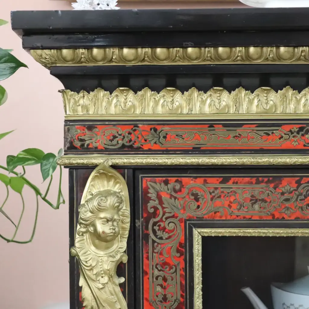 Vetrina alla Boulle in ebano, tartaruga e bronzi dorati Francia, seconda metà del XIX secolo – Stile Napoleone III Raffinata vetrina a un’anta realizzata secondo la celebre tecnica “alla Boulle”, con impiallacciatura in ebano e ricchi intarsi in ottone su fondo in tartaruga rossa naturale, disposti in eleganti motivi a racemi e volute di gusto Luigi XIV. Il prospetto è impreziosito da importanti applicazioni in bronzo dorato finemente cesellato, raffiguranti figure allegoriche laterali, fregi a foglie d’acanto e mascherone centrale nella parte inferiore. L’anta vetrata, incorniciata da profili dorati, apre su ripiani interni destinati all’esposizione di porcellane e oggetti d’arte. Esempio di alta ebanisteria francese del periodo Napoleone III, il mobile coniuga ricchezza ornamentale, equilibrio architettonico e forte impatto scenografico, risultando ideale per ambienti di rappresentanza o collezioni antiquarie di pregio.
