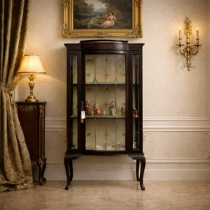 Vetrina inglese in mogano con vetri piombati, inizio XX secolo Elegante vetrina di manifattura inglese, databile ai primi anni del XX secolo, realizzata in mogano massello e impiallacciato, con struttura slanciata ed equilibrata. Il mobile poggia su gambe mosse a cabriole ed è caratterizzato da un’anta frontale centinata con vetri piombati artistici arricchiti da inserti verdi geometrici. I vetri laterali permettono una visione completa del contenuto, rendendola particolarmente adatta all’esposizione di porcellane, oggetti da collezione o cristalli. La fascia superiore è impreziosita da delicati intarsi floreali, mentre l’interno, rivestito in carta decorata, è dotato di ripiani in vetro che valorizzano la presentazione degli oggetti. Il mogano conserva una bella patina calda, con lievi segni del tempo coerenti con l’età. Vetrina raffinata e versatile, ideale per ambienti classici, studi o salotti.