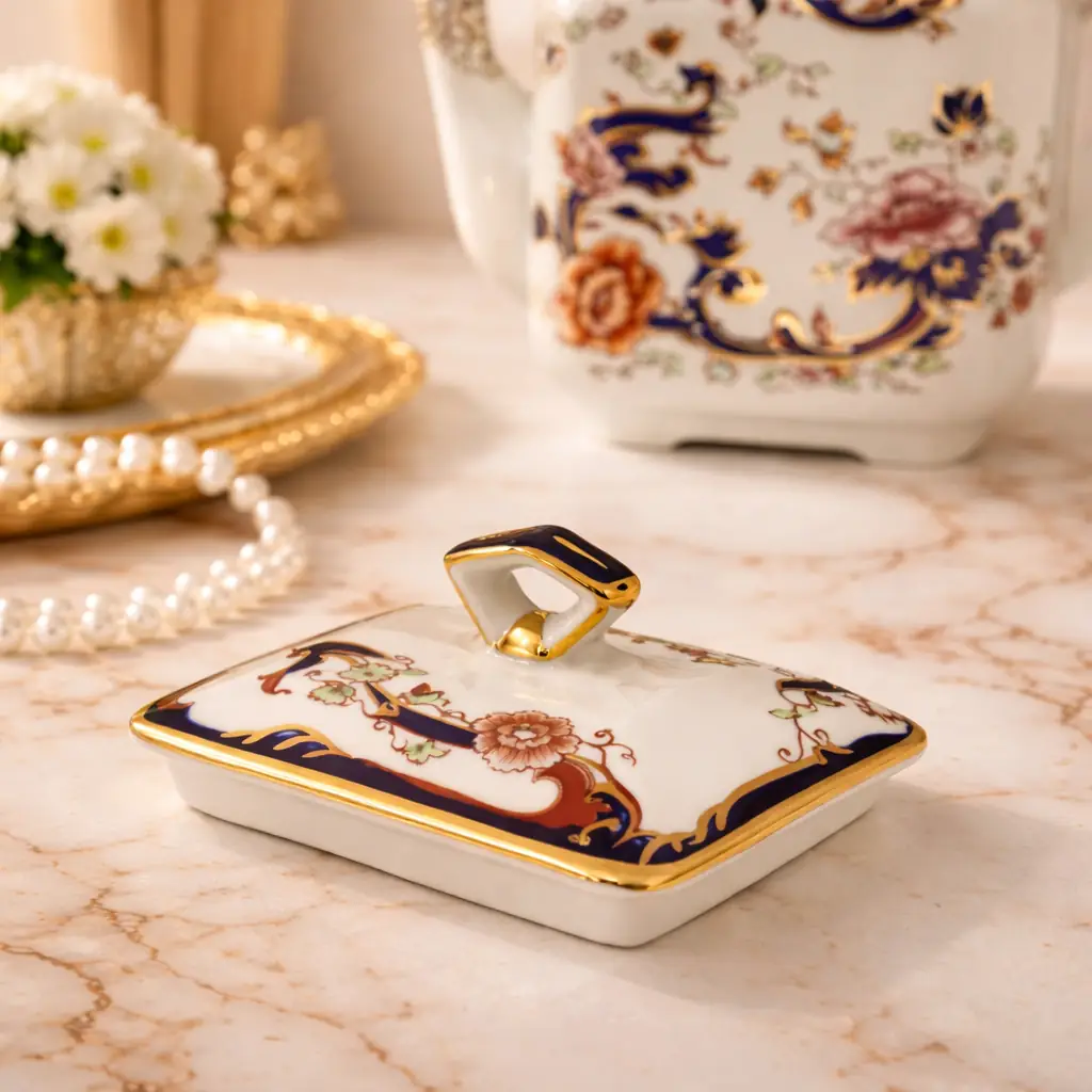 Teiera in porcellana Mason’s – Decoro “Mandalay” Raffinata teiera in porcellana inglese Mason’s “Patent Ironstone”, decorata nel celebre motivo “Mandalay”, di ispirazione orientale. Il fondo avorio è arricchito da un elegante intreccio di peonie e racemi policromi nei toni del rosa, ocra e verde, incorniciati da sinuose volute in blu cobalto intenso finemente profilate in oro. La forma sfaccettata e architettonica, con beccuccio slanciato e manico superiore sagomato con inserti blu e dorati, conferisce all’insieme grande equilibrio e presenza scenica. Il coperchio, perfettamente combaciante, riprende la medesima ricchezza decorativa. Al verso reca il marchio con corona “Mason’s Patent Ironstone – Mandalay – Made in England”. Ottimo stato di conservazione, con smalti brillanti e dorature ben preservate. Ideale sia per collezionisti di porcellana inglese sia come elegante complemento d’arredo.