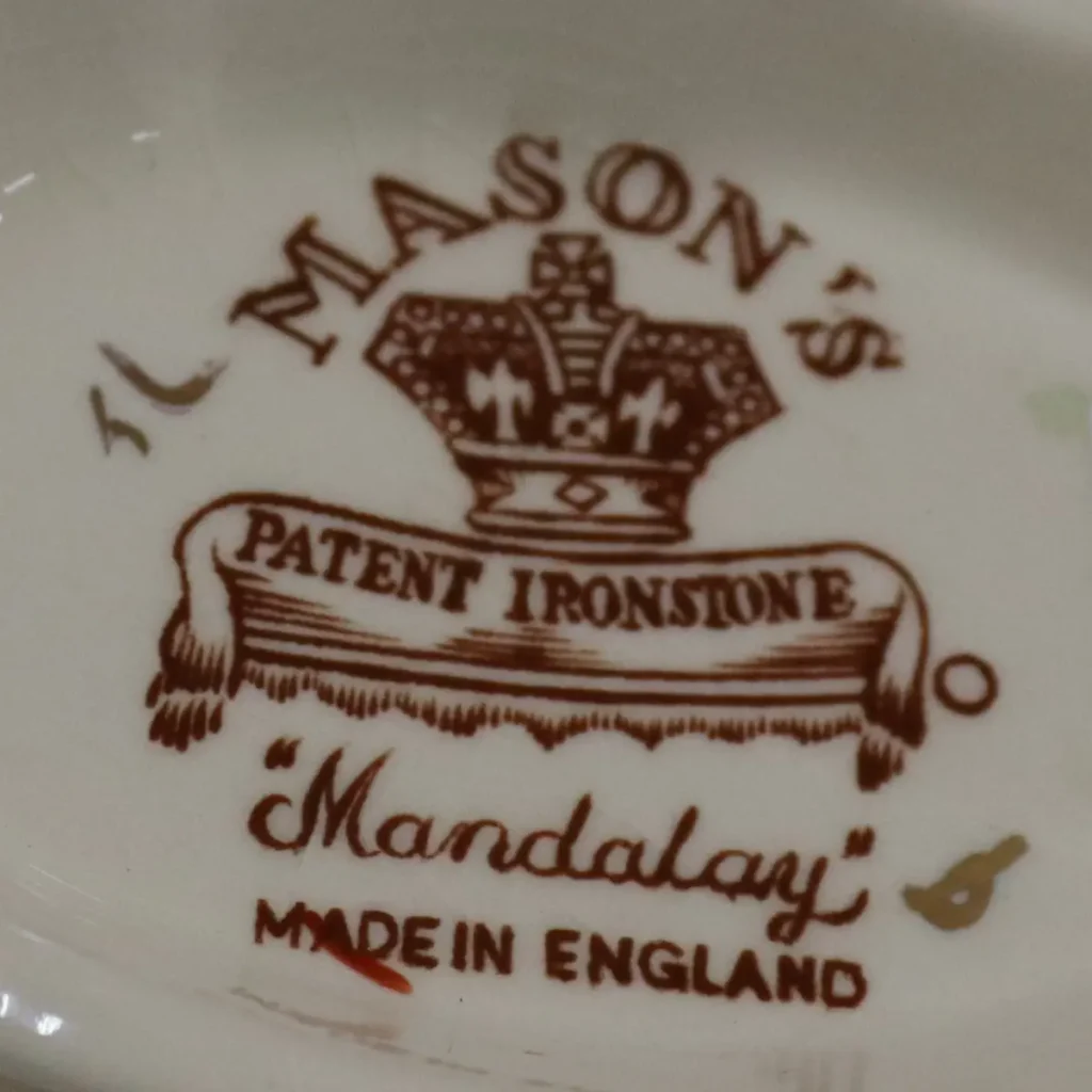 Teiera in porcellana Mason’s – Decoro “Mandalay” Raffinata teiera in porcellana inglese Mason’s “Patent Ironstone”, decorata nel celebre motivo “Mandalay”, di ispirazione orientale. Il fondo avorio è arricchito da un elegante intreccio di peonie e racemi policromi nei toni del rosa, ocra e verde, incorniciati da sinuose volute in blu cobalto intenso finemente profilate in oro. La forma sfaccettata e architettonica, con beccuccio slanciato e manico superiore sagomato con inserti blu e dorati, conferisce all’insieme grande equilibrio e presenza scenica. Il coperchio, perfettamente combaciante, riprende la medesima ricchezza decorativa. Al verso reca il marchio con corona “Mason’s Patent Ironstone – Mandalay – Made in England”. Ottimo stato di conservazione, con smalti brillanti e dorature ben preservate. Ideale sia per collezionisti di porcellana inglese sia come elegante complemento d’arredo.