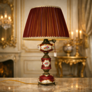 Lampada da tavolo in porcellana dipinta e bronzo, Francia, fine XIX – inizi XX secolo Elegante lampada da tavolo di manifattura francese, realizzata tra la fine del XIX e i primi anni del XX secolo. Il fusto è composto da elementi in porcellana smaltata e dipinta a mano, decorati con riserve floreali policrome su fondo rosso porpora, impreziosite da filettature dorate. La struttura è completata da una montatura in bronzo cesellato, con base sagomata su piedini e braccio laterale con chiave di accensione e tirante in cordoncino. La lampada è dotata di paralume tronco-conico in tessuto plissettato, rifinito con passamaneria decorativa, armonizzato per colore e stile con il corpo della lampada. Oggetto di gusto tardo ottocentesco, decorativo e funzionale, si presenta in ottimo stato di conservazione, con lievi segni del tempo coerenti con l’età.