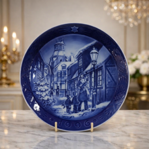Piatto da collezione in porcellana della storica manifattura danese Royal Copenhagen, edizione annuale 1996. Decorato nel tradizionale blu cobalto su fondo bianco, raffigura una suggestiva scena invernale di villaggio con figure e architetture d’epoca, resa con grande finezza grafica e profondità tonale. La tesa presenta eleganti motivi ornamentali in rilievo che incorniciano la composizione centrale, esaltando la qualità della smaltatura e la cura esecutiva tipica della produzione Royal Copenhagen. Pensato come oggetto decorativo da parete o da esposizione su supporto, rappresenta un classico intramontabile del collezionismo di porcellane nord-europee del secondo Novecento.