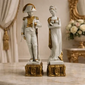 Coppia di figure in porcellana di Capodimonte firmate “Merli” – soggetto napoleonico Elegante coppia di sculture in porcellana di Capodimonte, finemente modellate e decorate a mano con applicazioni in oro, raffiguranti Napoleone Bonaparte in uniforme con bicorno e dama in abito stile Impero. Le figure, di gusto neoclassico, presentano una buona qualità esecutiva nei dettagli dei volti e nei panneggi, con smaltatura lucida e dorature che valorizzano l’insieme. Entrambe poggiano su basi architettoniche riccamente decorate con motivi a rilievo e medaglione con iniziale “N”, chiaro richiamo all’iconografia napoleonica. Marchio sottosmalto della N coronata di Capodimonte e firma incisa “Merli”. Produzione italiana della seconda metà del ’900. Buono stato generale, con lievi segni del tempo e minime usure alle dorature. Ideali come oggetti decorativi o da collezione.