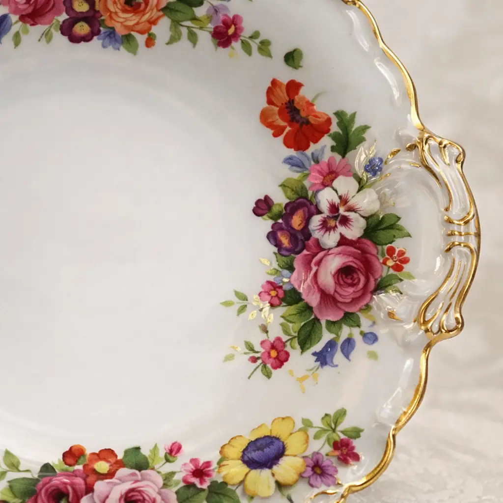 Piatto da dolce in porcellana europea, con bordo sagomato e anse laterali stilizzate. Decorato a mazzi floreali policromi disposti a ghirlanda, con profilature in oro lungo il bordo e sulle prese. Produzione della prima metà del XX secolo. In ottime condizioni generali. Elegante oggetto da tavola e da collezione.