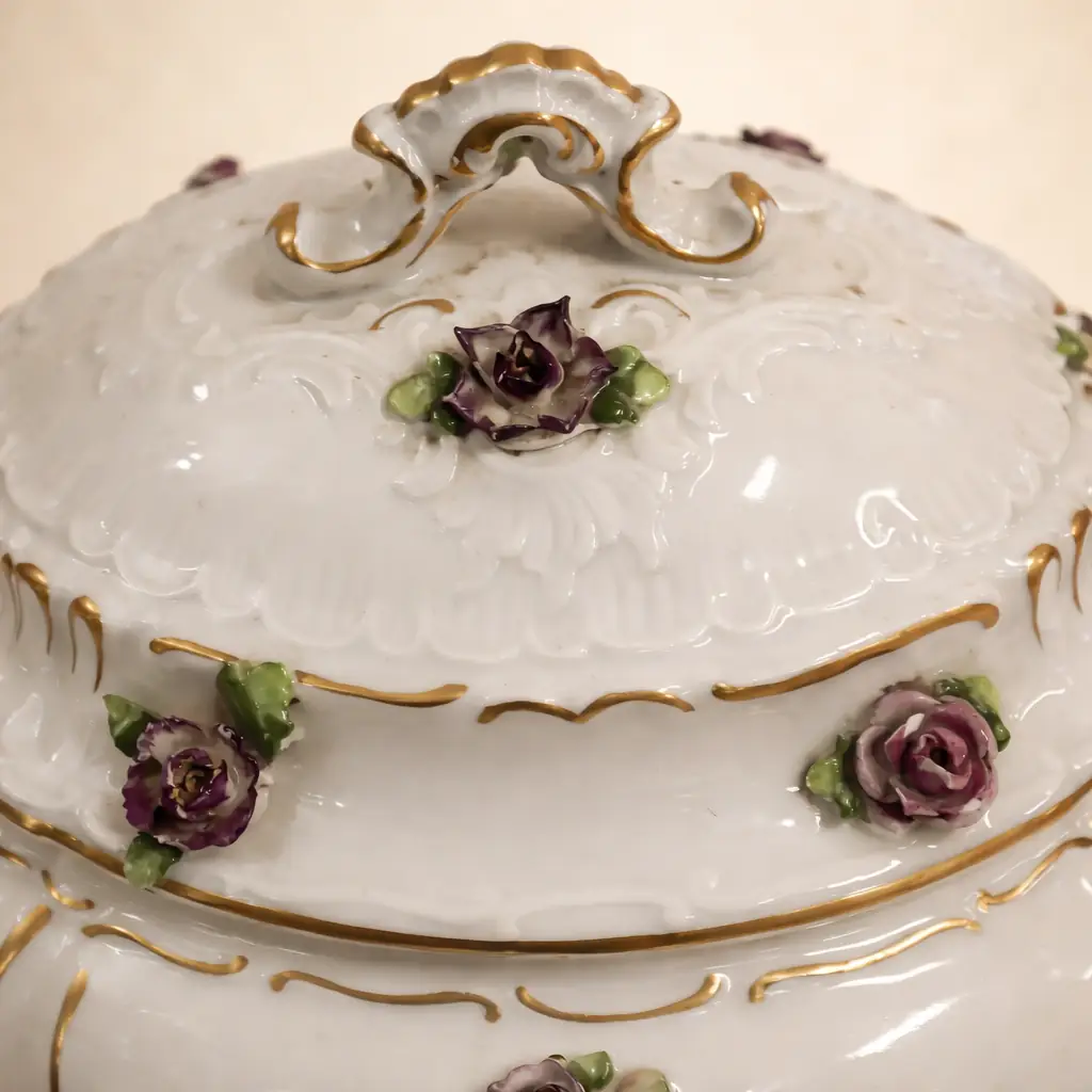 Zuppiera in porcellana decorata a rilievo con applicazioni floreali Manifattura europea, fine XIX – inizi XX secolo Raffinata zuppiera in porcellana bianca modellata, impreziosita da eleganti motivi rocaille in bassorilievo e ricca decorazione floreale plastica applicata a mano, raffigurante rose policrome e piccoli fiori dai toni vivaci. I profili e le volute sono rifiniti in oro, che valorizza la morbida modellazione del corpo bombato e del coperchio sagomato con presa dorata. Il corpo poggia su quattro piedini ricurvi anch’essi decorati in oro, conferendo slancio ed equilibrio all’insieme. Al verso è presente marchio in blu con “N” sormontata da corona, riconducibile alla produzione di capodimonte tra la fine dell’Ottocento e i primi decenni del Novecento. Buono stato di conservazione generale, con lievi segni del tempo e leggere usure alla doratura coerenti con l’epoca. Elegante oggetto da collezione e complemento d’arredo per ambienti classici o eclettici.