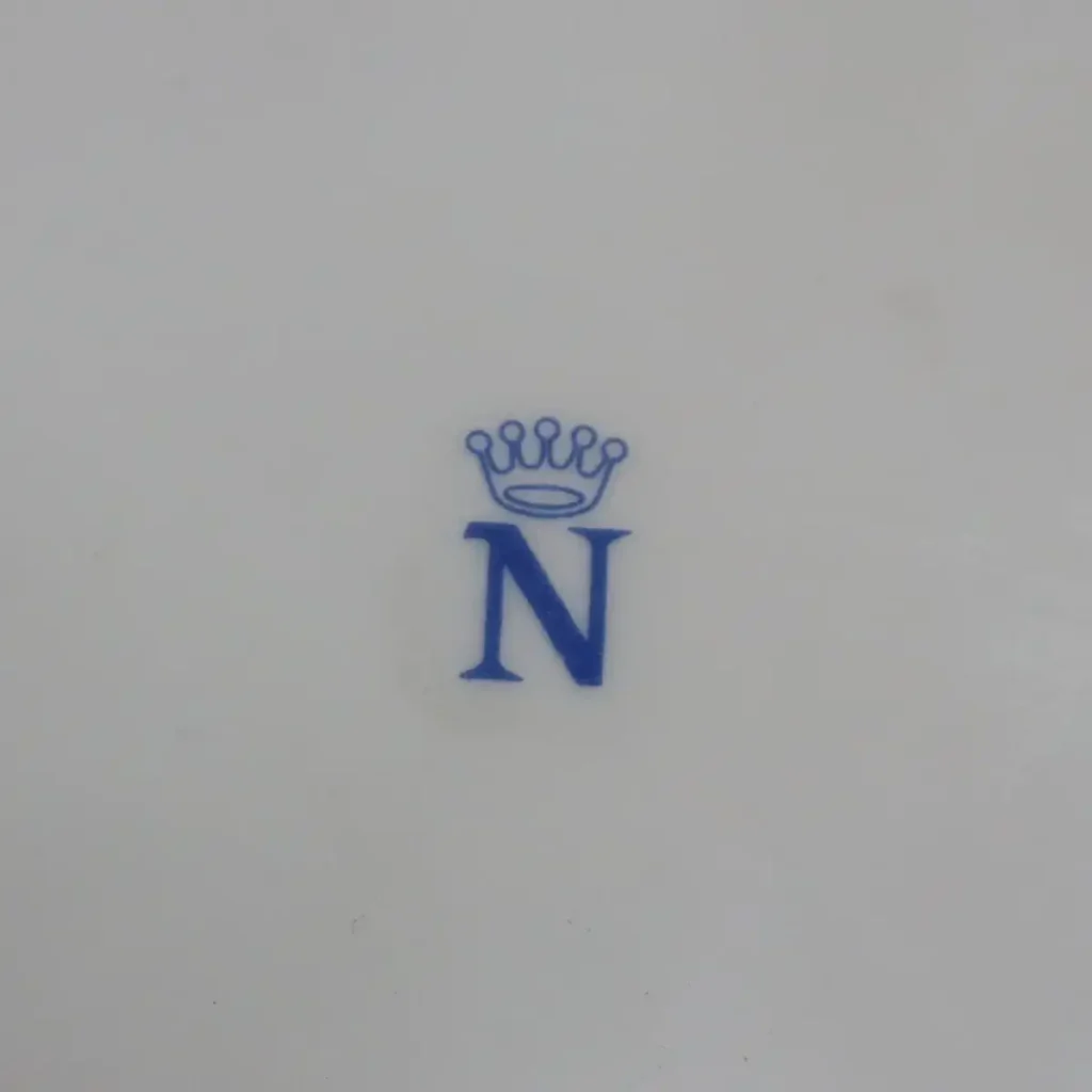 Zuppiera in porcellana decorata a rilievo con applicazioni floreali Manifattura europea, fine XIX – inizi XX secolo Raffinata zuppiera in porcellana bianca modellata, impreziosita da eleganti motivi rocaille in bassorilievo e ricca decorazione floreale plastica applicata a mano, raffigurante rose policrome e piccoli fiori dai toni vivaci. I profili e le volute sono rifiniti in oro, che valorizza la morbida modellazione del corpo bombato e del coperchio sagomato con presa dorata. Il corpo poggia su quattro piedini ricurvi anch’essi decorati in oro, conferendo slancio ed equilibrio all’insieme. Al verso è presente marchio in blu con “N” sormontata da corona, riconducibile alla produzione di capodimonte tra la fine dell’Ottocento e i primi decenni del Novecento. Buono stato di conservazione generale, con lievi segni del tempo e leggere usure alla doratura coerenti con l’epoca. Elegante oggetto da collezione e complemento d’arredo per ambienti classici o eclettici.